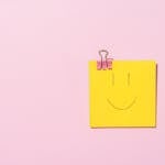 Yellow sticky note with a smiley face clipped to a pink surface, conveying happiness and simplicity.