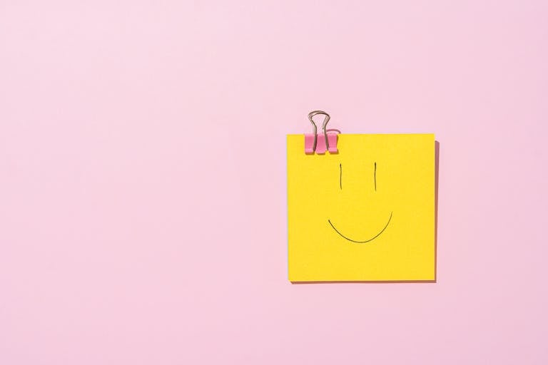 Yellow sticky note with a smiley face clipped to a pink surface, conveying happiness and simplicity.